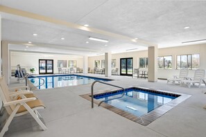 Une piscine intérieure avec un bord carrelé, des chaises longues et une vue sur l’extérieur grâce à de grandes fenêtres.