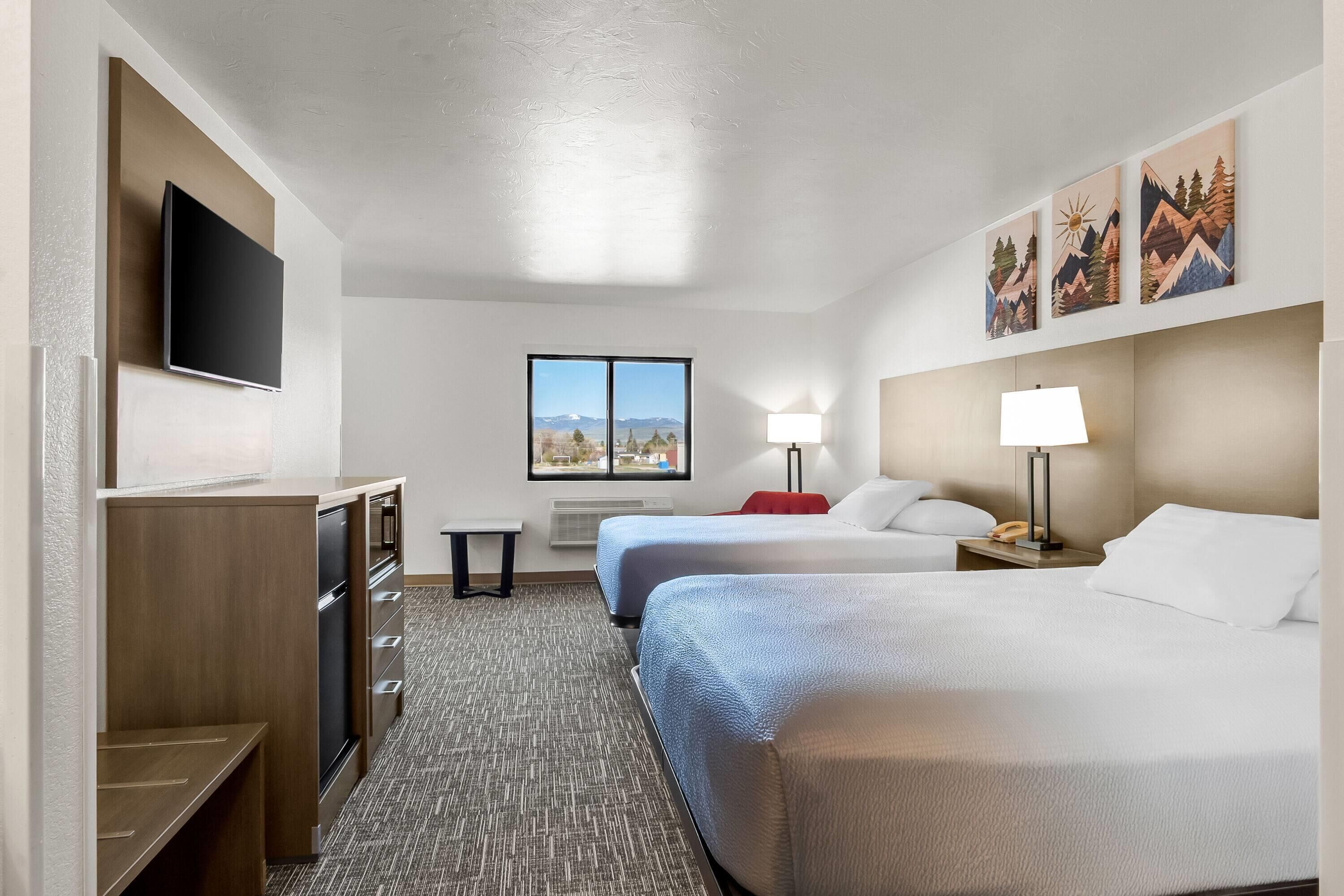 A hotel room with two beds, a TV, a window with a mountain view, and artwork on the wall.