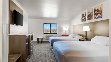 A hotel room with two beds, a TV, a window with a mountain view, and artwork on the wall.
