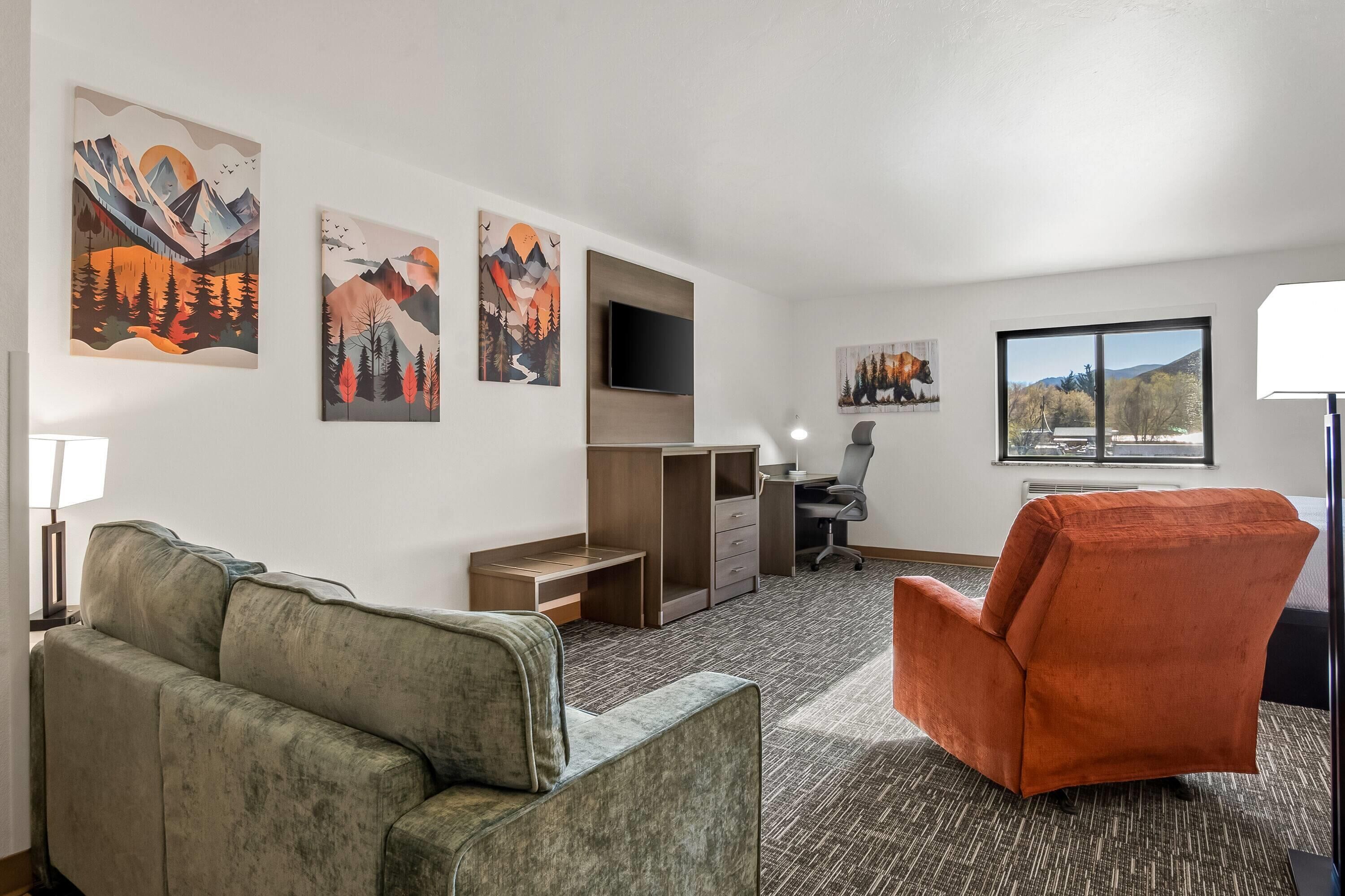 A hotel room with a sofa, an orange armchair, a desk with a chair, a flat-screen TV, and artwork on the walls.