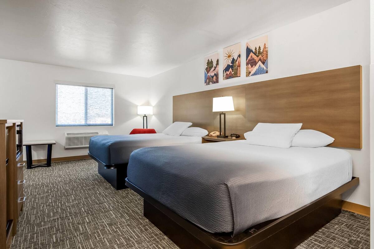 Habitación de hotel con dos camas, un escritorio, una silla, un ventanal y cuadros en la pared.