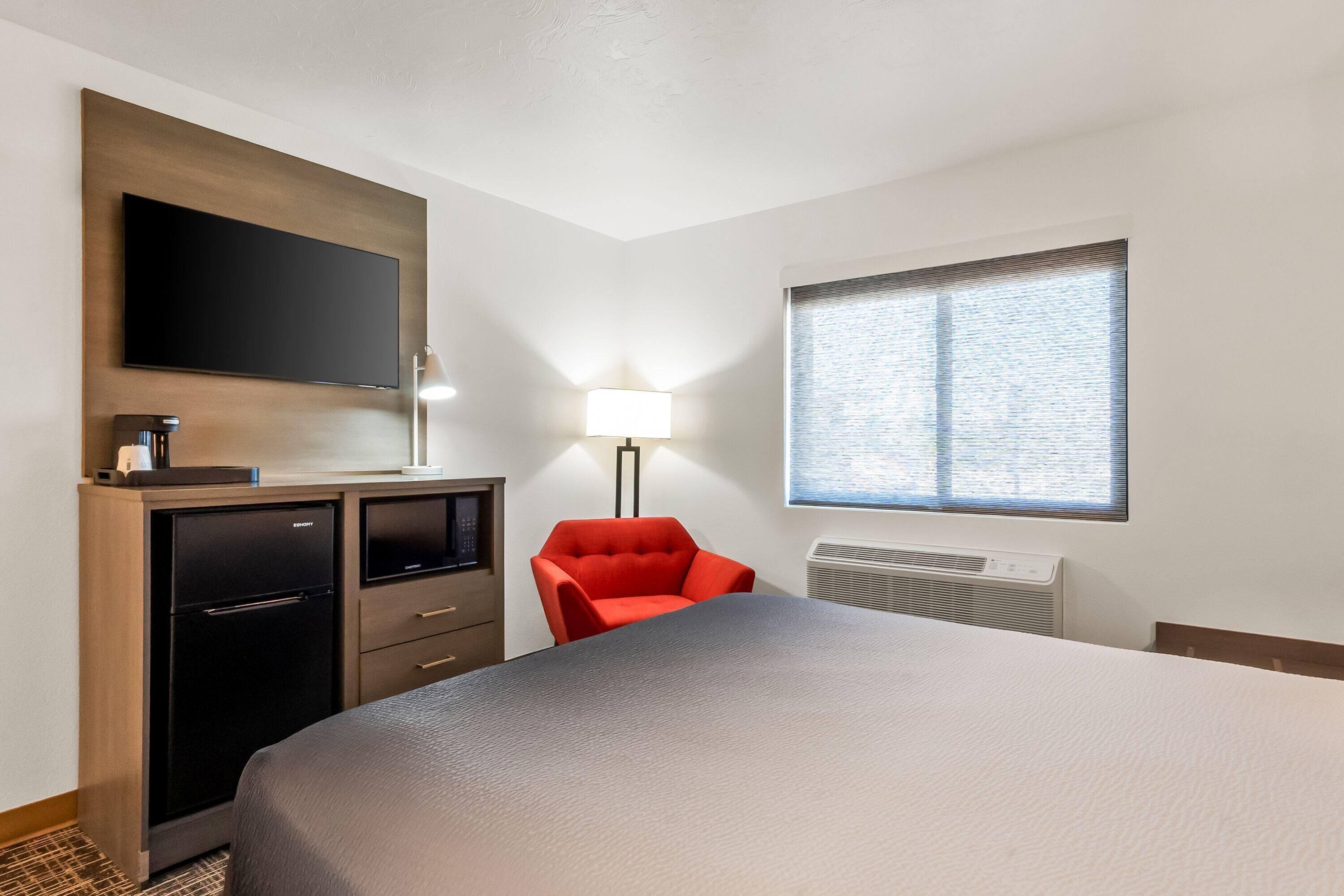 Habitación de hotel con cama, escritorio, televisor de pantalla plana, una silla roja, lámpara y ventana con persianas.