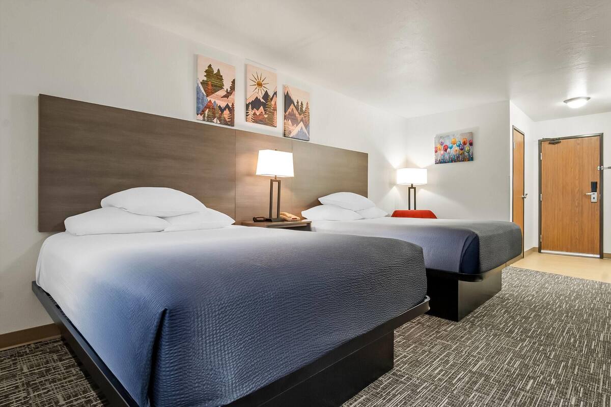 Habitación de hotel con dos camas, cabecera de madera, mesita de noche con lámpara y cuadros en las paredes.