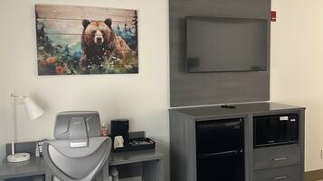 A hotel room with a desk, chair, bed, TV, and a bear painting on the wall.