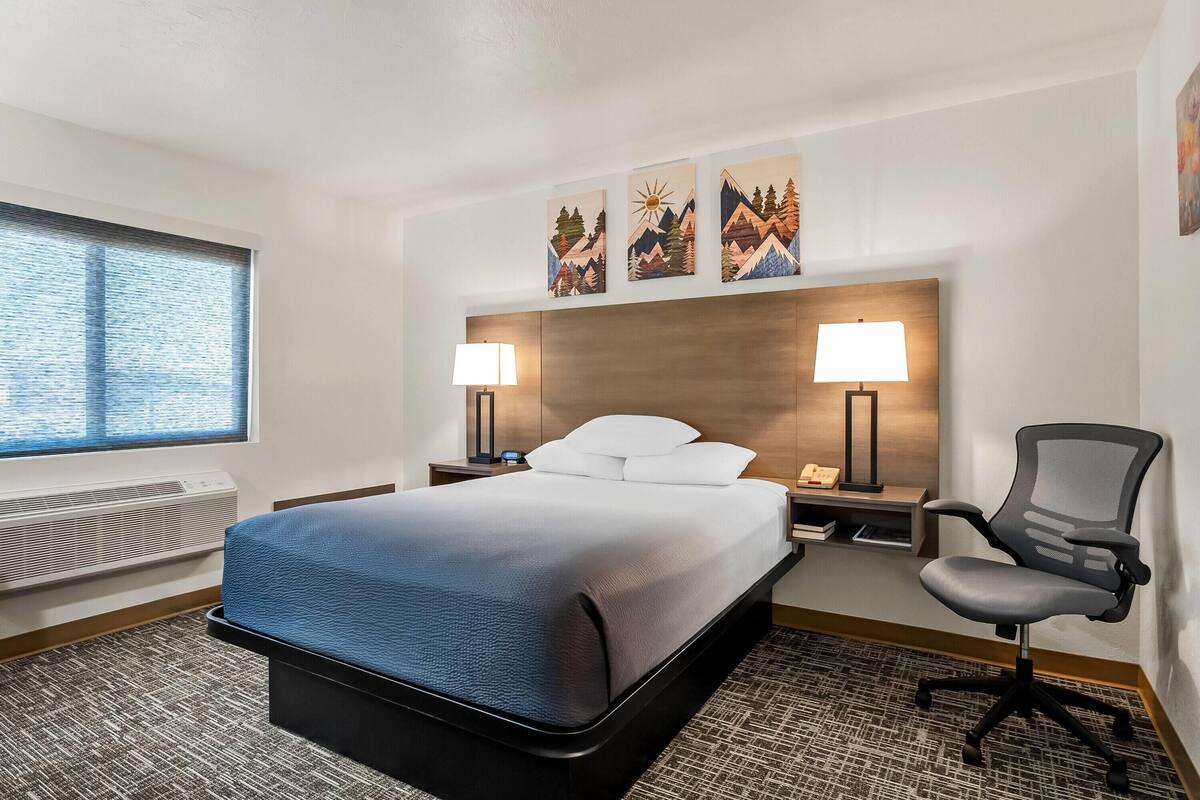 Habitación de hotel con una cama, dos lámparas de noche, un escritorio, una silla y tres cuadros colgados en la pared.