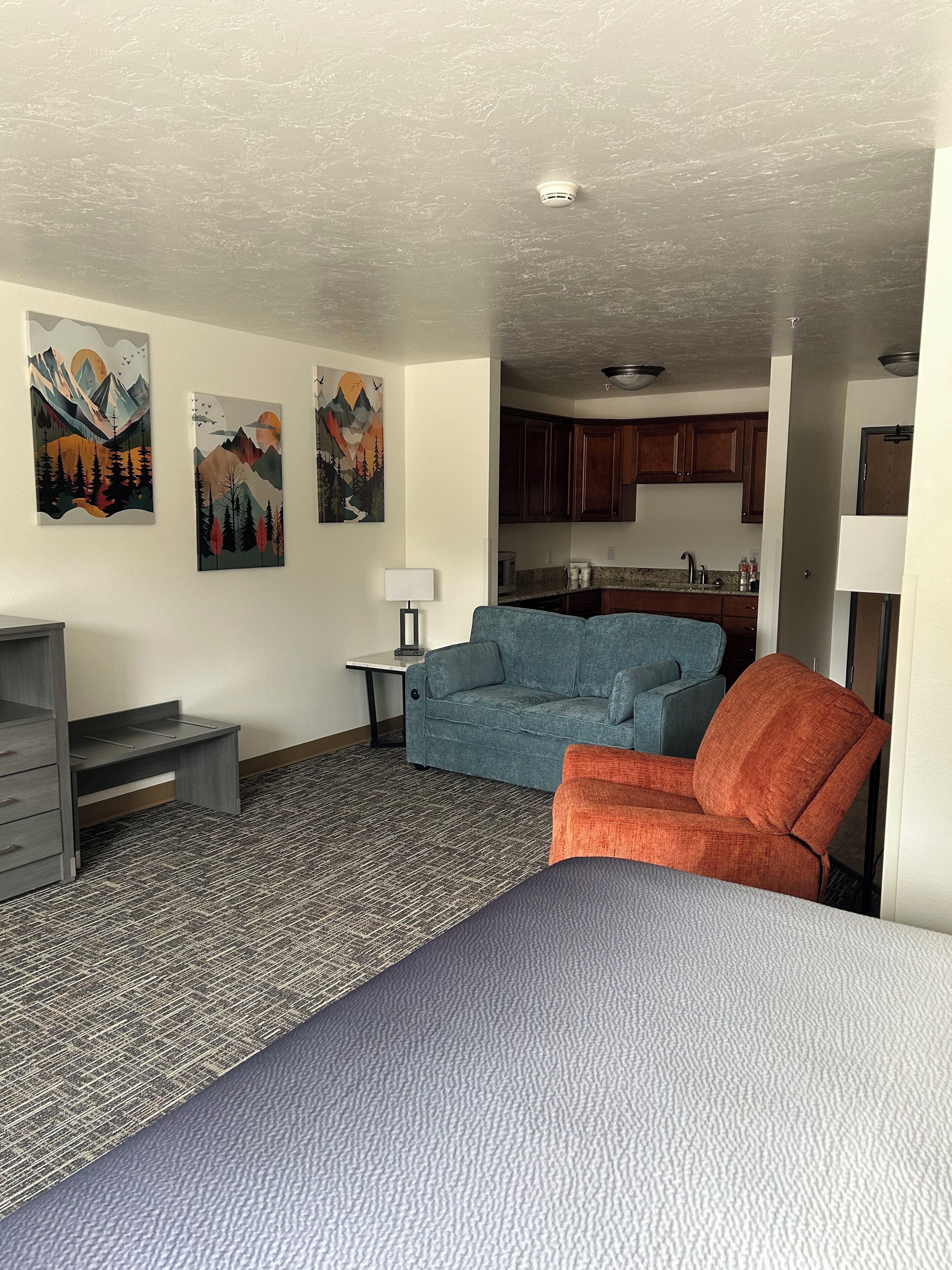 A compact room with a bed, a blue sofa, an orange armchair, a desk, and a kitchenette in the background.