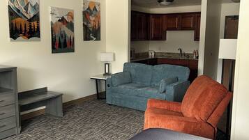 A compact room with a bed, a blue sofa, an orange armchair, a desk, and a kitchenette in the background.