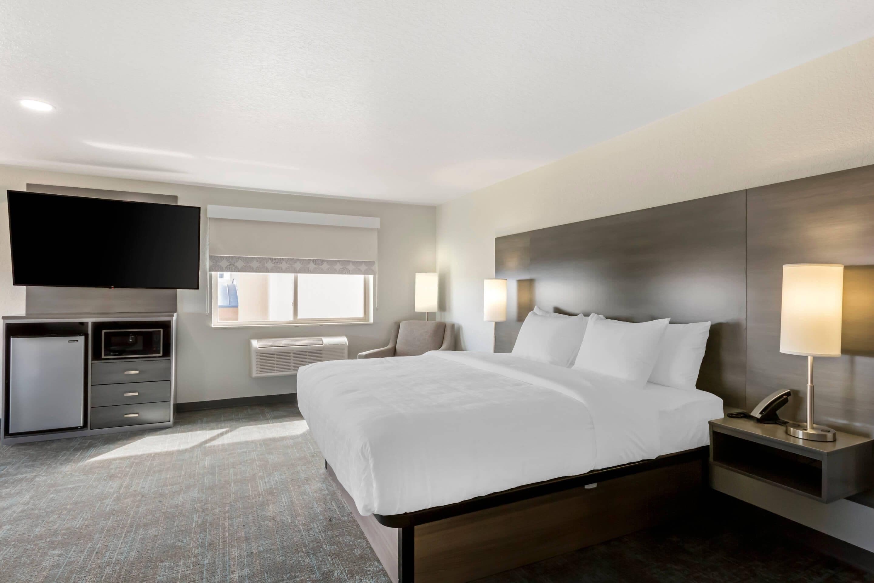 Quarto de hotel com uma cama grande, televisão de tela plana, escrivaninha com abajur, mini-geladeira e janela com persianas.