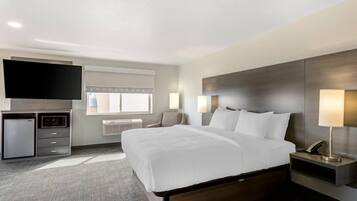 Quarto de hotel com uma cama grande, televisão de tela plana, escrivaninha com abajur, mini-geladeira e janela com persianas.