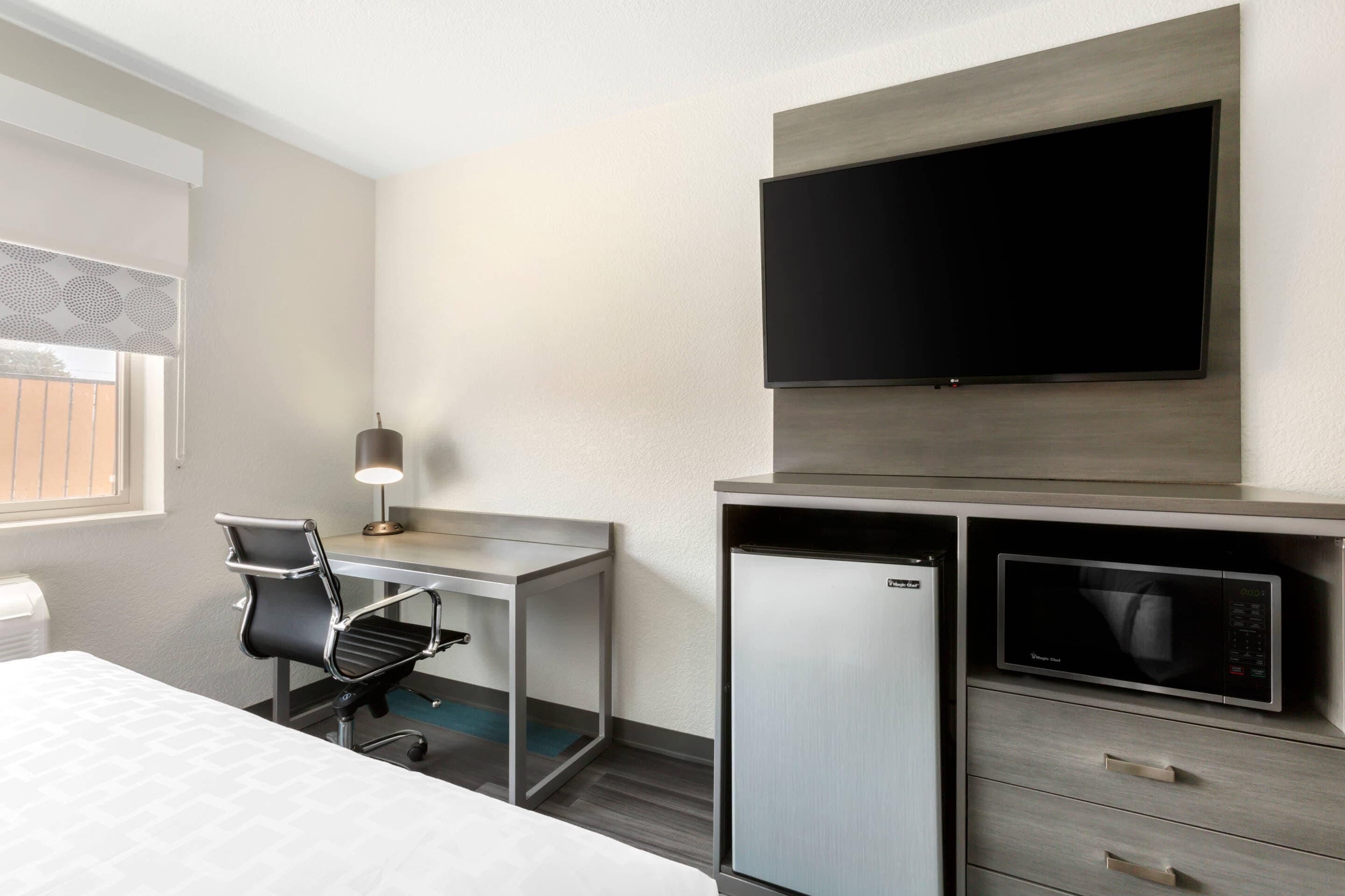 A hotel room with a bed, a desk with a chair, a flat-screen TV mounted on the wall, a microwave, and a mini-fridge.