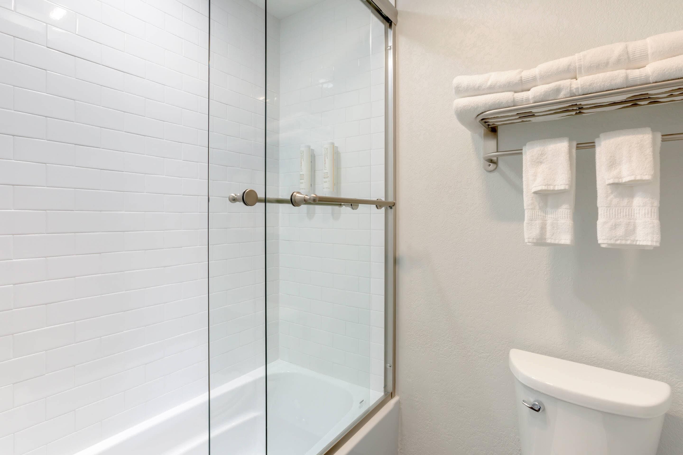 A bathroom with a glass-enclosed shower, white tiled walls, a white toilet, and a towel rack with multiple towels.