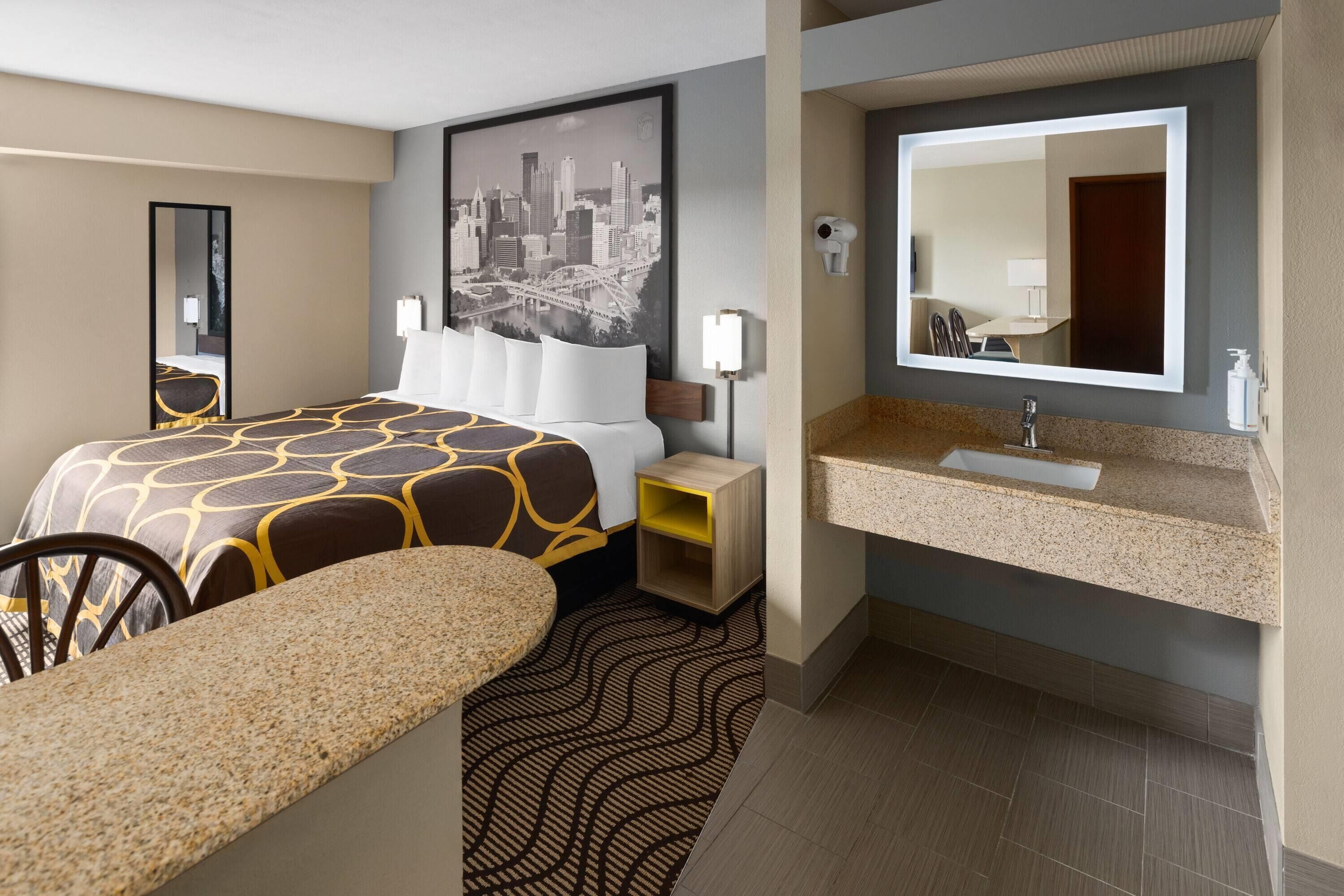 Habitación de hotel con cama, escritorio, silla, baño con lavamanos y espejo, y un cuadro con vista a la ciudad.