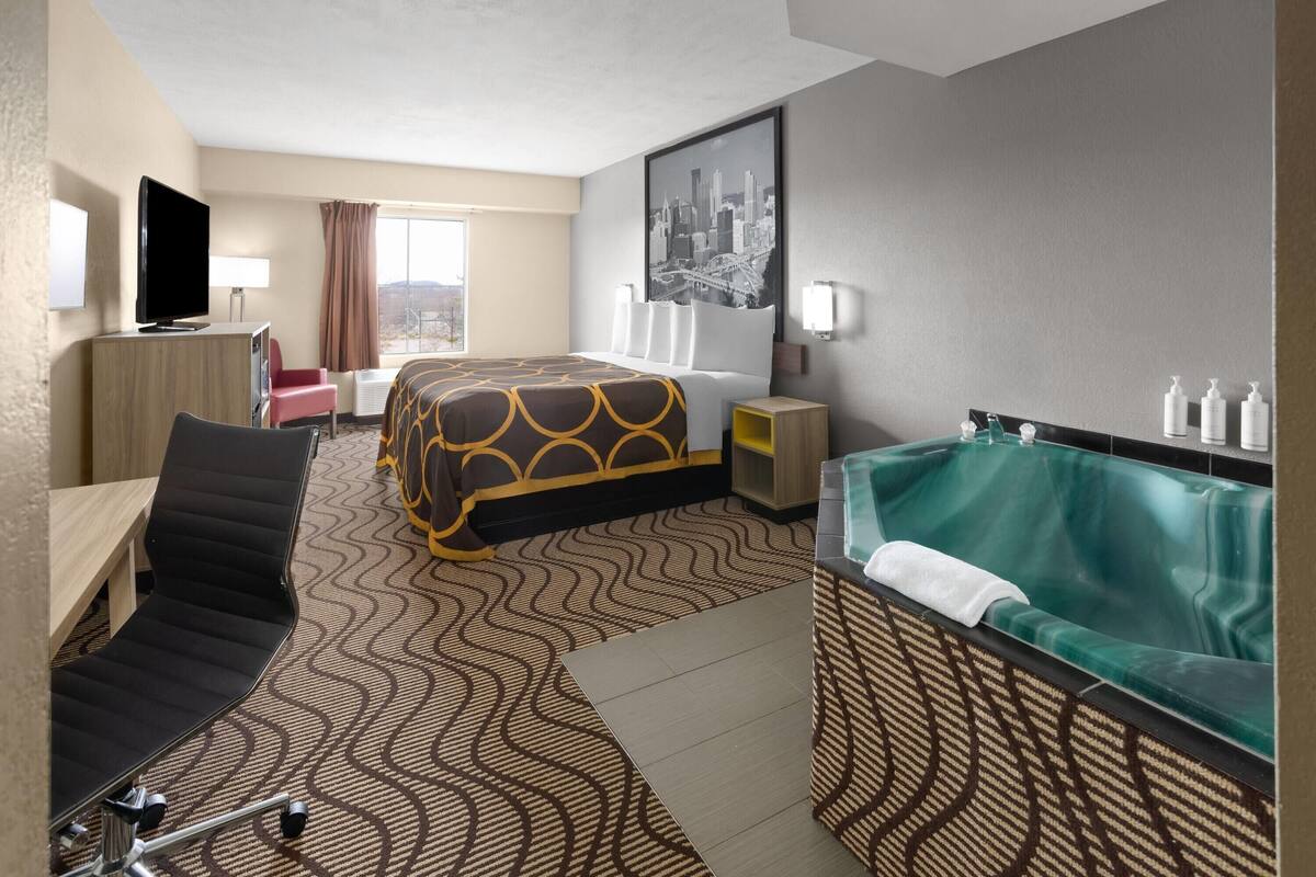 Hotelkamer met een bed, bureau, stoel, televisie, raam met gordijnen, wandlamp, stadsgezicht in een lijst en een bad.