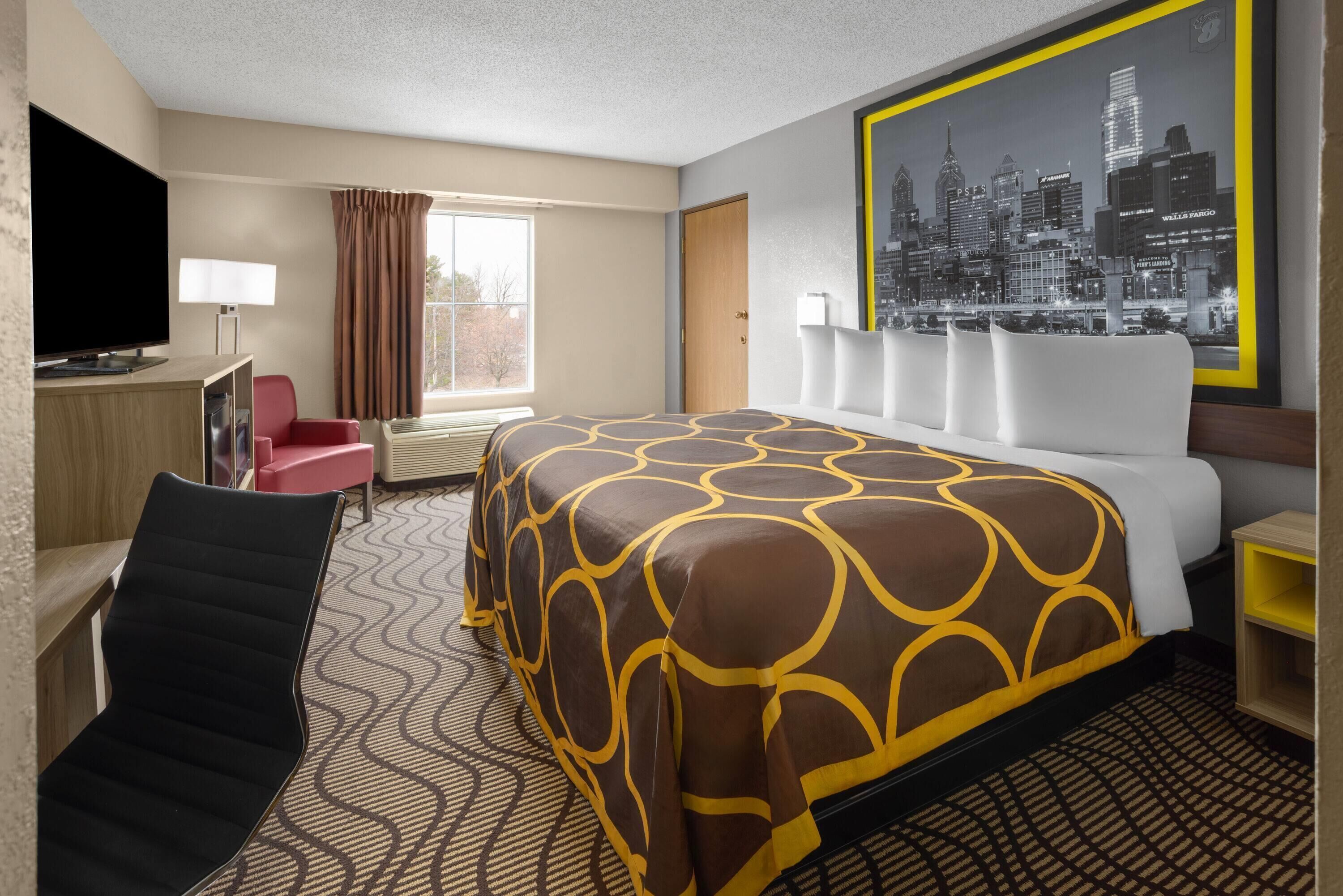 A hotel room with a bed, a television, a desk, a chair, a window with curtains, and a cityscape poster on the wall.