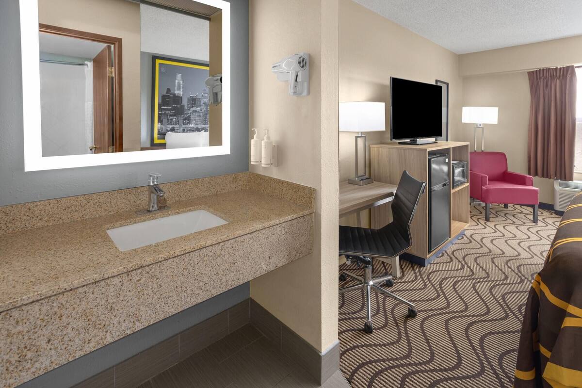 Hotelkamer met een wastafel met granieten blad, een flatscreen televisie, een bureau met stoel, een rode fauteuil en een grote spiegel.