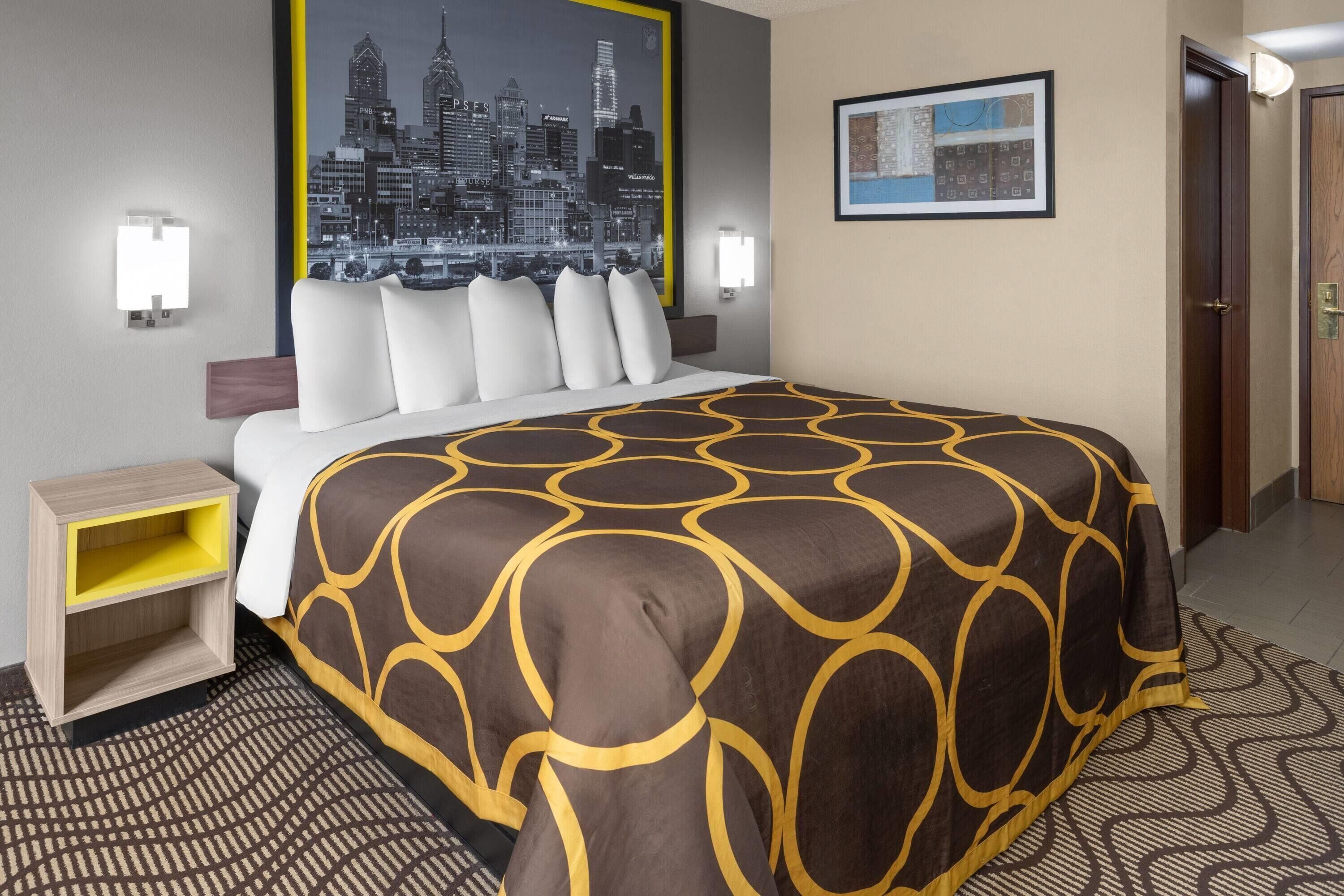 Ein Hotelzimmer mit Bett, Nachttisch, einem Stadtbild-Poster, einem gerahmten Bild und einer Tür.