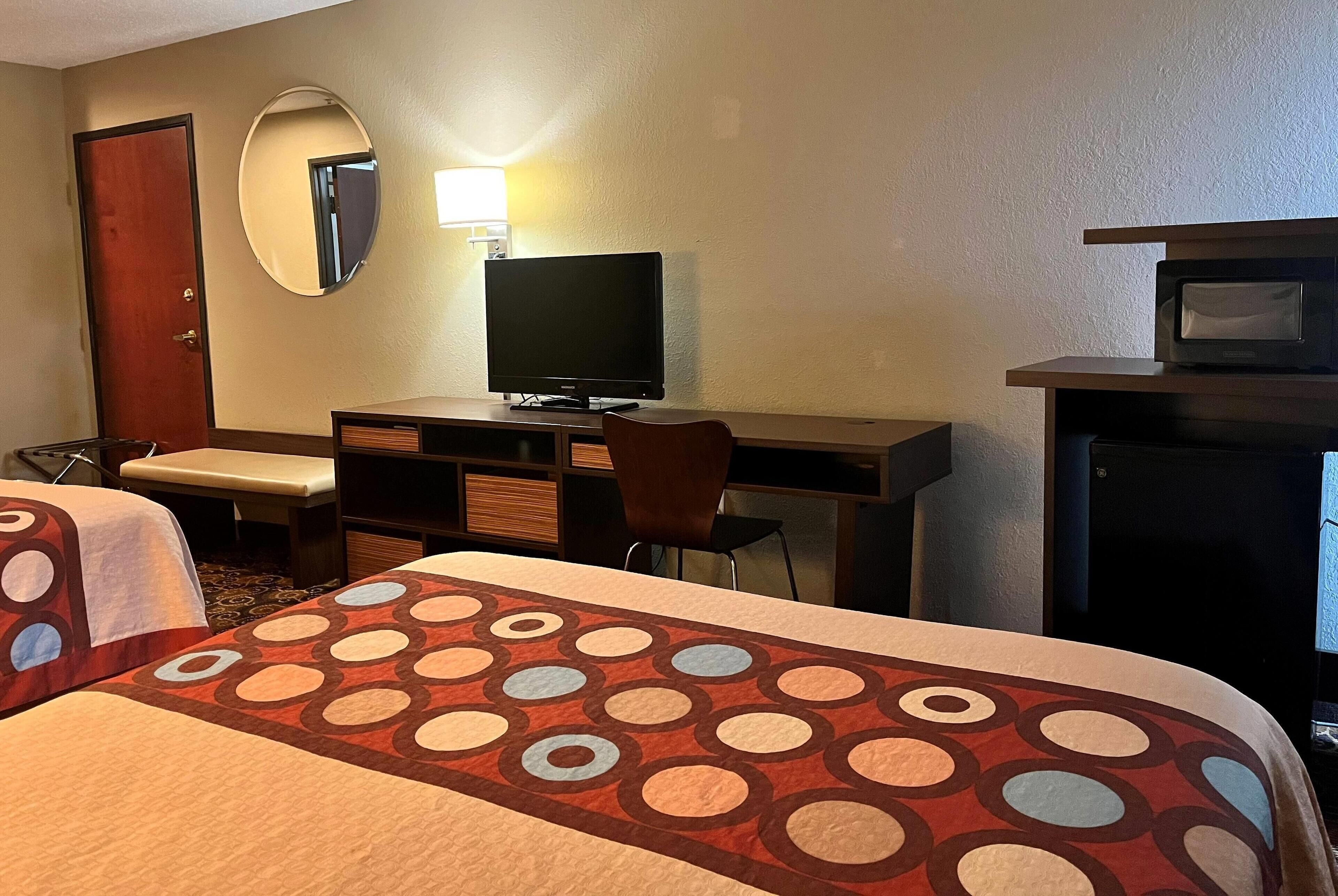 Quarto de hotel com duas camas, uma escrivaninha com televisão, uma cadeira, uma luminária de mesa e um micro-ondas.