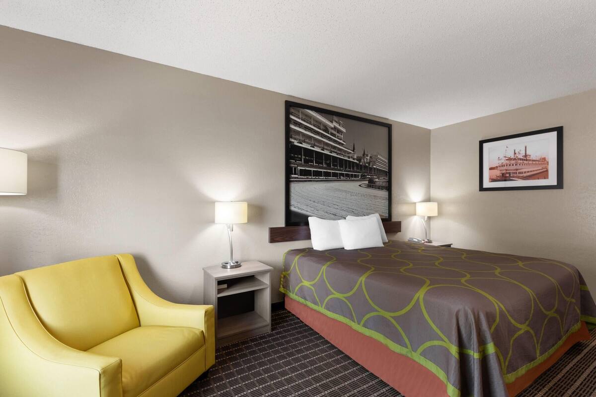Una habitación de hotel con una cama, un sillón amarillo, una mesita de noche con lámpara y dos cuadros en la pared.