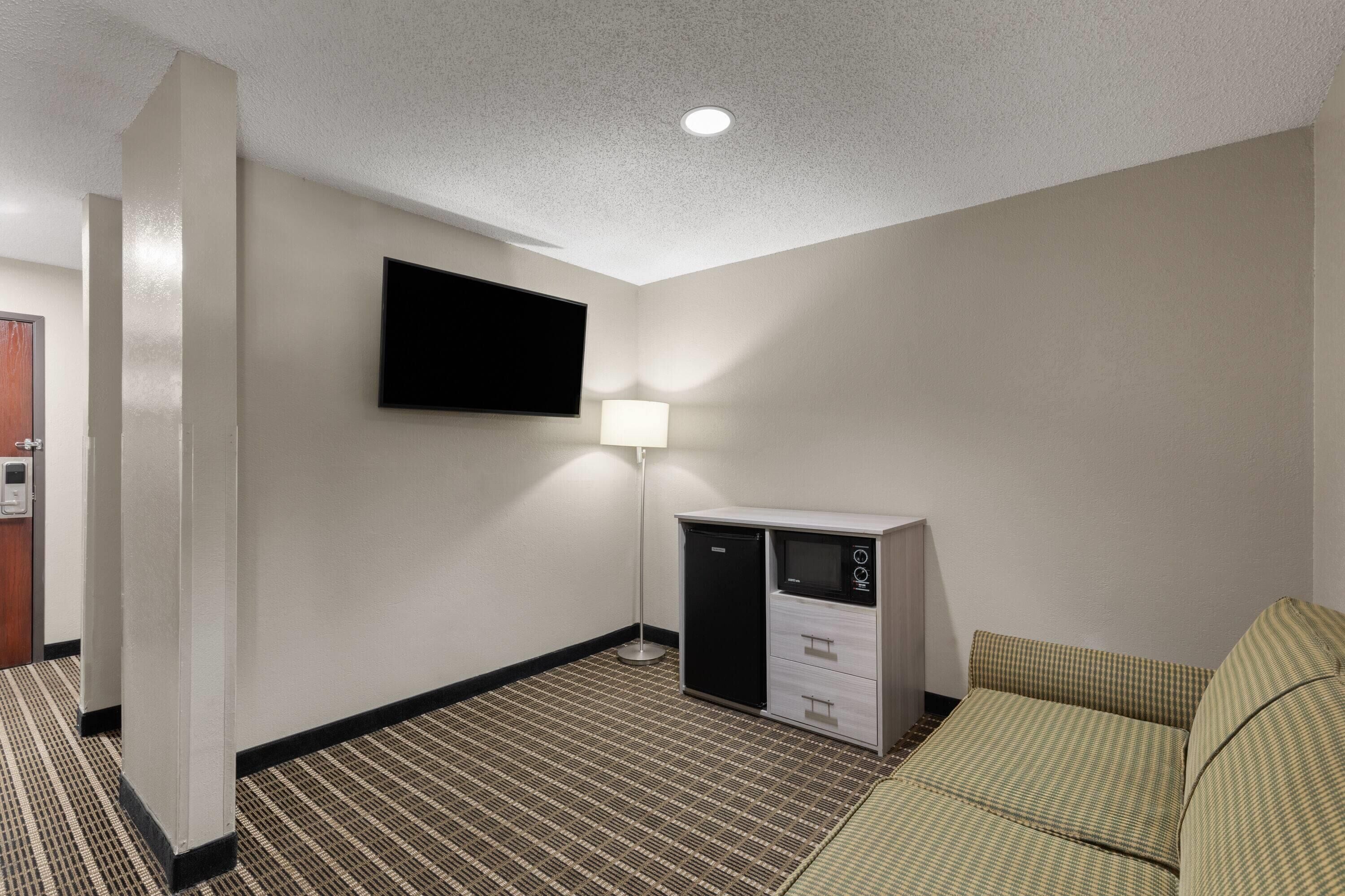 A hotel room with a flat-screen TV mounted on the wall, a desk with a lamp, a refrigerator, and a patterned carpet.