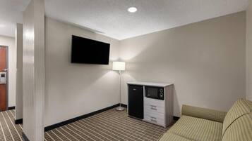A hotel room with a flat-screen TV mounted on the wall, a desk with a lamp, a refrigerator, and a patterned carpet.
