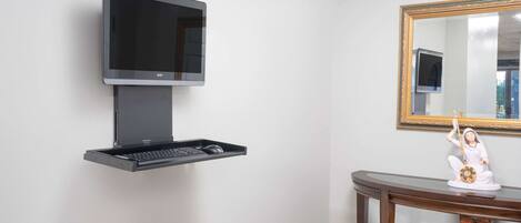 A wall-mounted television with a keyboard tray below it, a wooden console table with a statue, and a mirror with a gold frame.