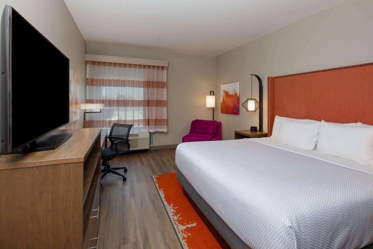 A hotel room with a large bed, a desk with a flat-screen TV, a chair, and a window with curtains.