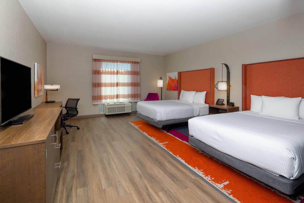 A hotel room with two beds, a desk with a flat-screen TV, a chair, a window with curtains, and a nightstand with a lamp.