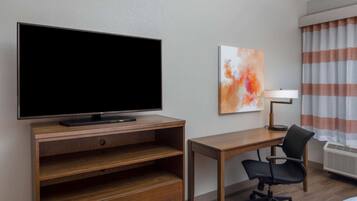 A hotel room with a flat-screen TV on a wooden stand, a desk with a chair, a painting on the wall, and a window with striped curtains.