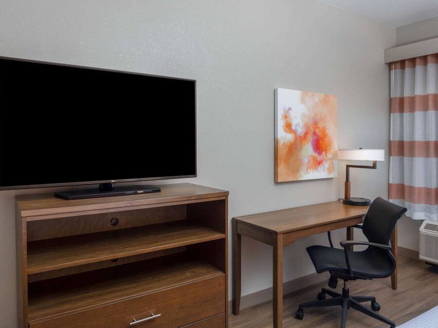 A hotel room with a flat-screen TV on a wooden stand, a desk with a chair, a painting on the wall, and a window with striped curtains.