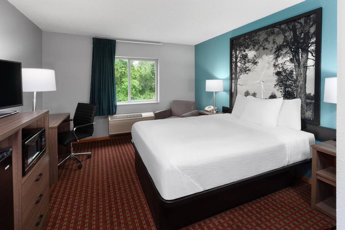 Quarto de hotel com cama, escrivaninha, cadeira, televisão e janela com cortinas.
