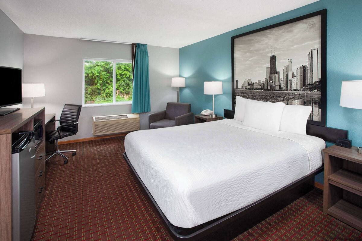 Quarto de hotel com uma cama grande, uma escrivaninha com computador, uma cadeira, uma televisão e uma janela com cortinas.
