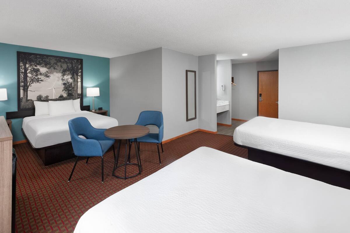 Quarto de hotel com duas camas, uma escrivaninha, duas cadeiras, uma mesinha e um quadro na parede.
