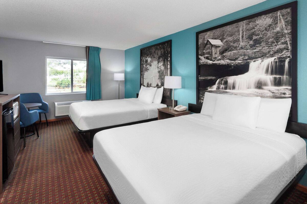 Quarto de hotel com duas camas, uma escrivaninha com cadeira, uma janela com cortinas e um grande painel na parede retratando uma cachoeira.