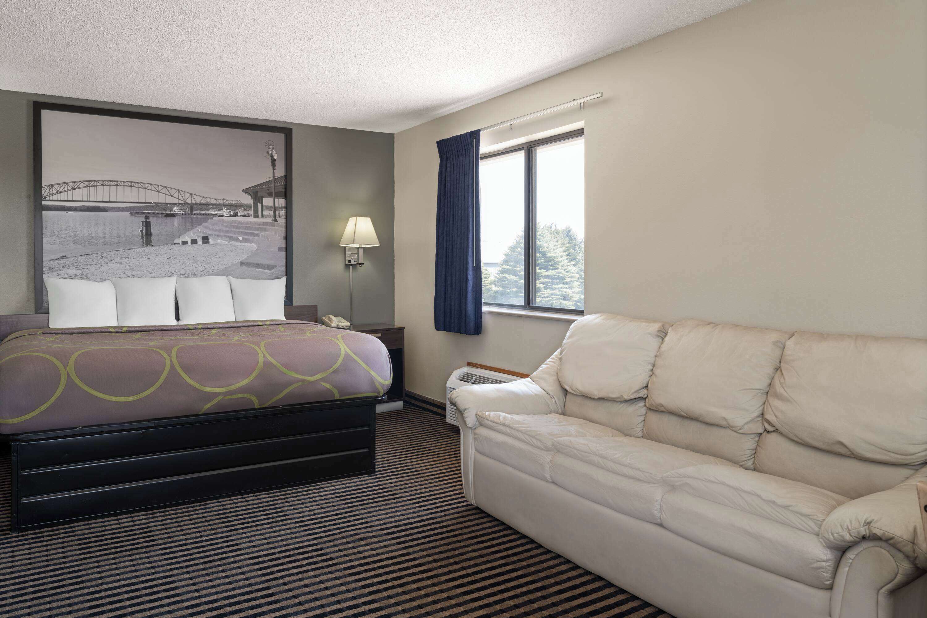 Habitación de hotel con cama, sofá, ventana grande, lámpara y aire acondicionado instalado en la pared.