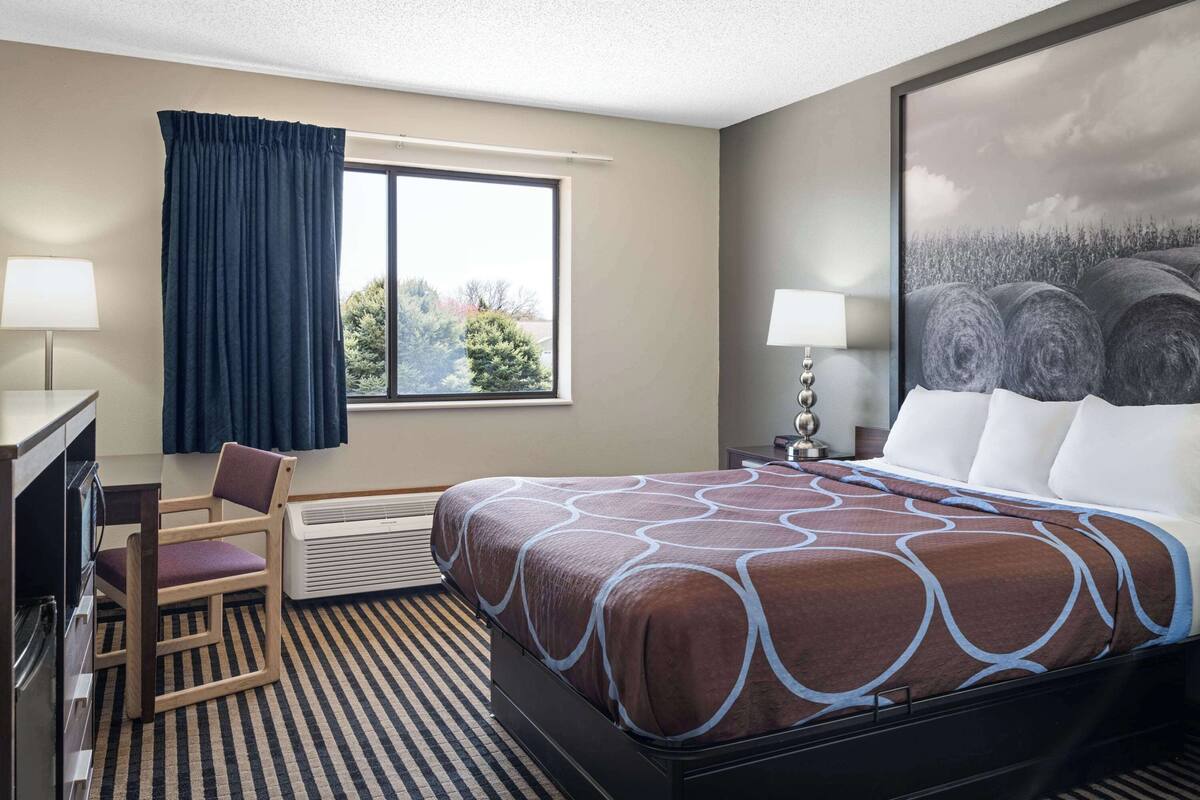 A hotel room with a bed, a desk, a chair, a window with curtains, and a painting on the wall.