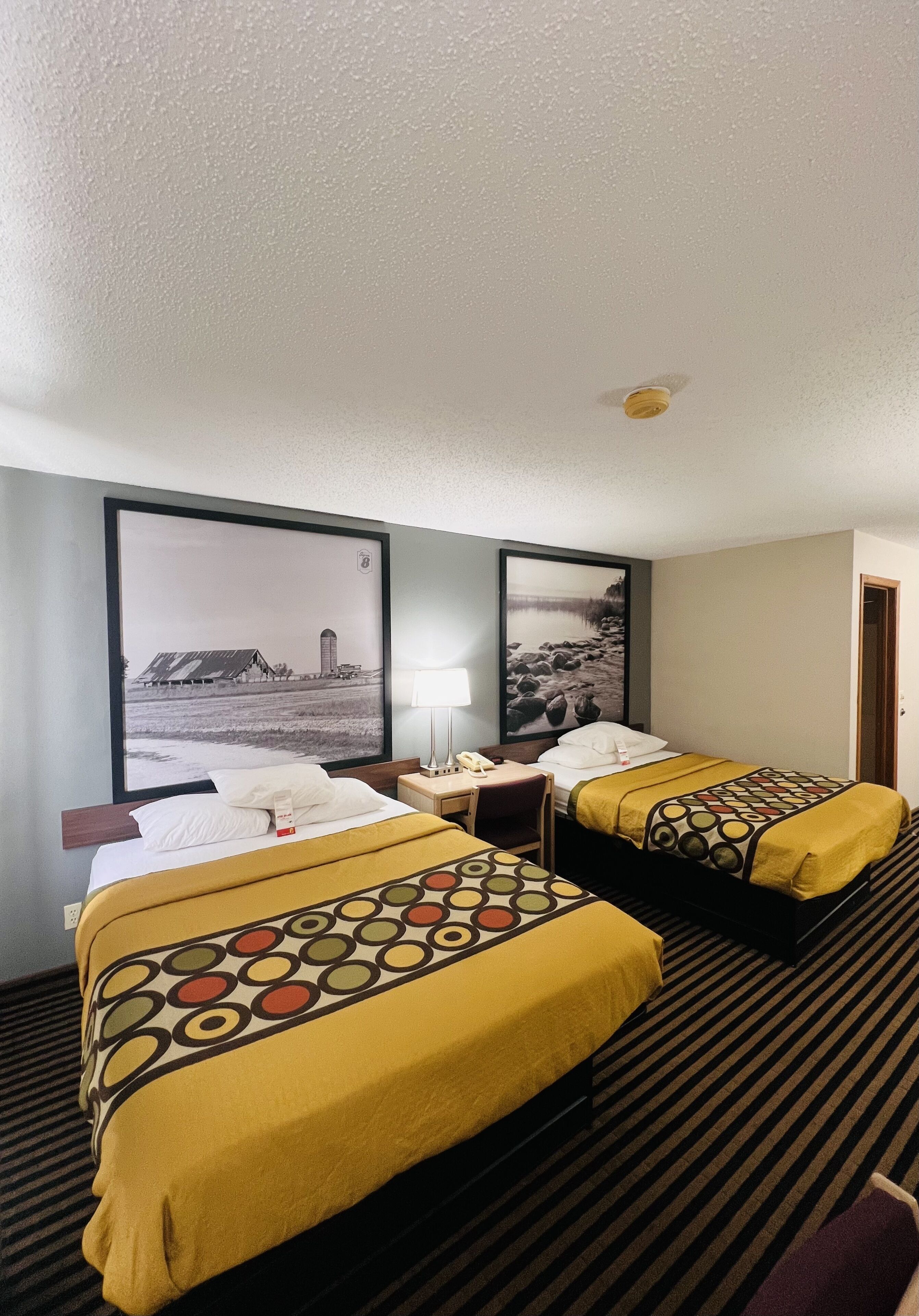 A hotel room with two beds, a desk, and a chair. There are two large framed pictures on the wall.