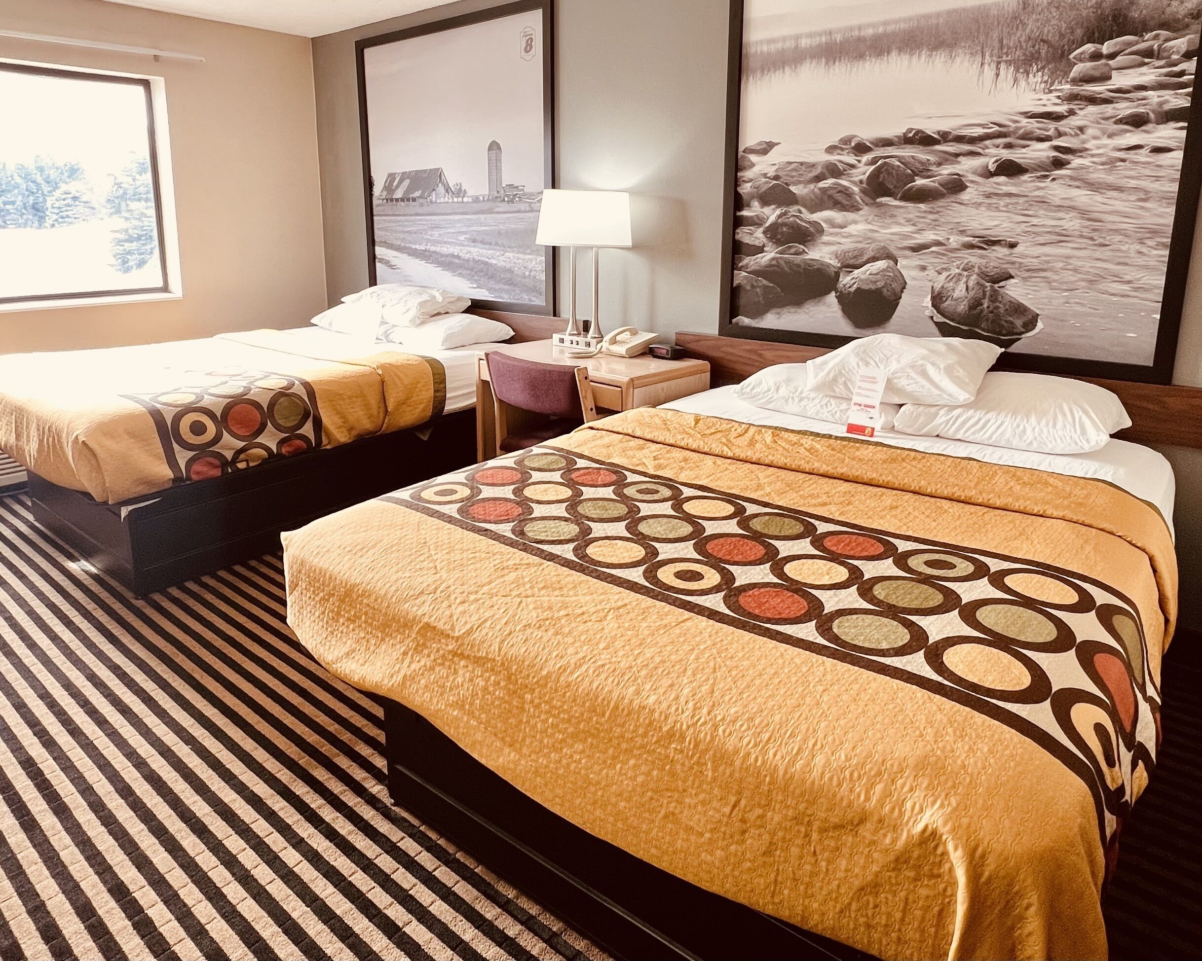 A hotel room with two beds, a bedside table with a lamp, and a large framed picture on the wall.
