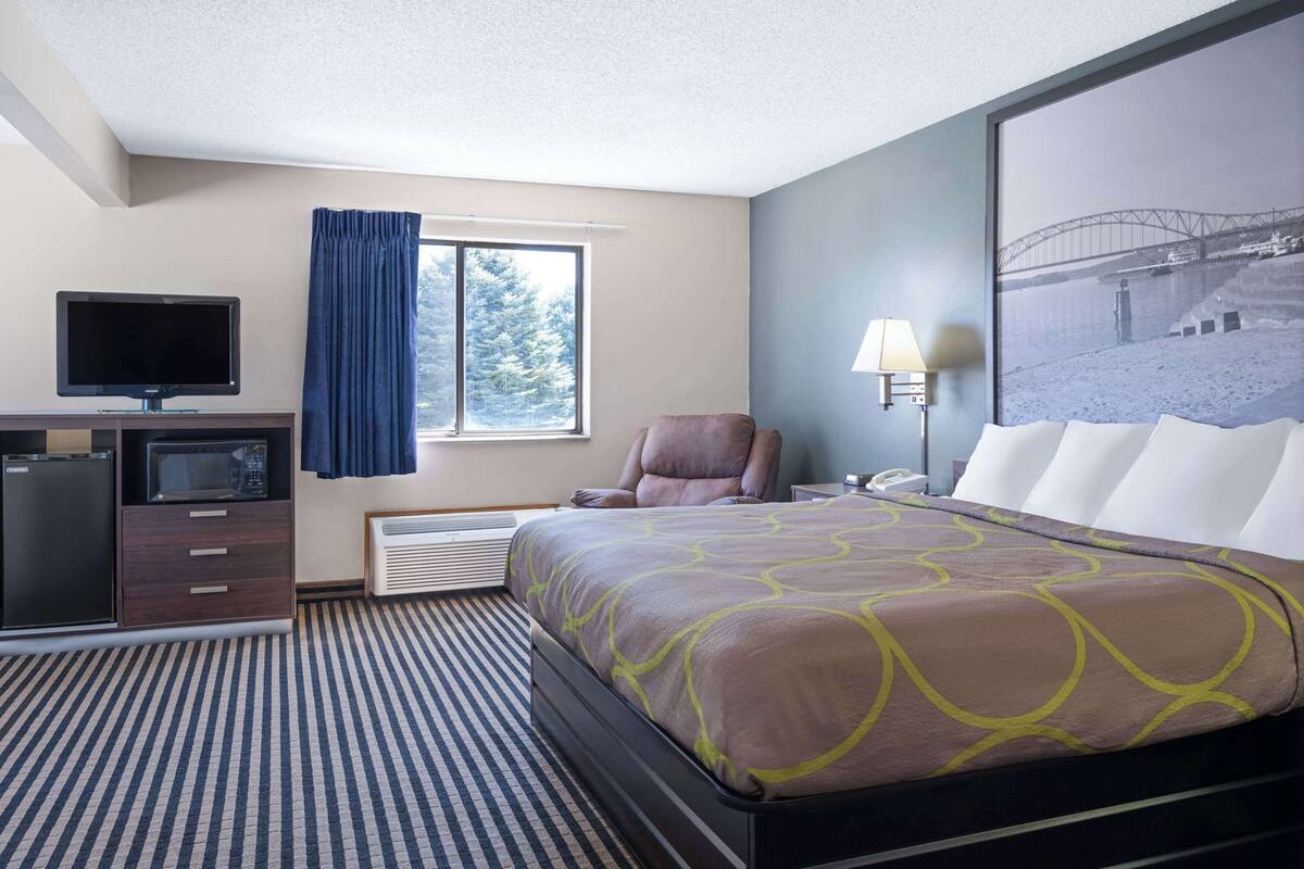 A hotel room with a bed, a desk with a TV, a chair, a window with curtains, and a wall-mounted lamp.