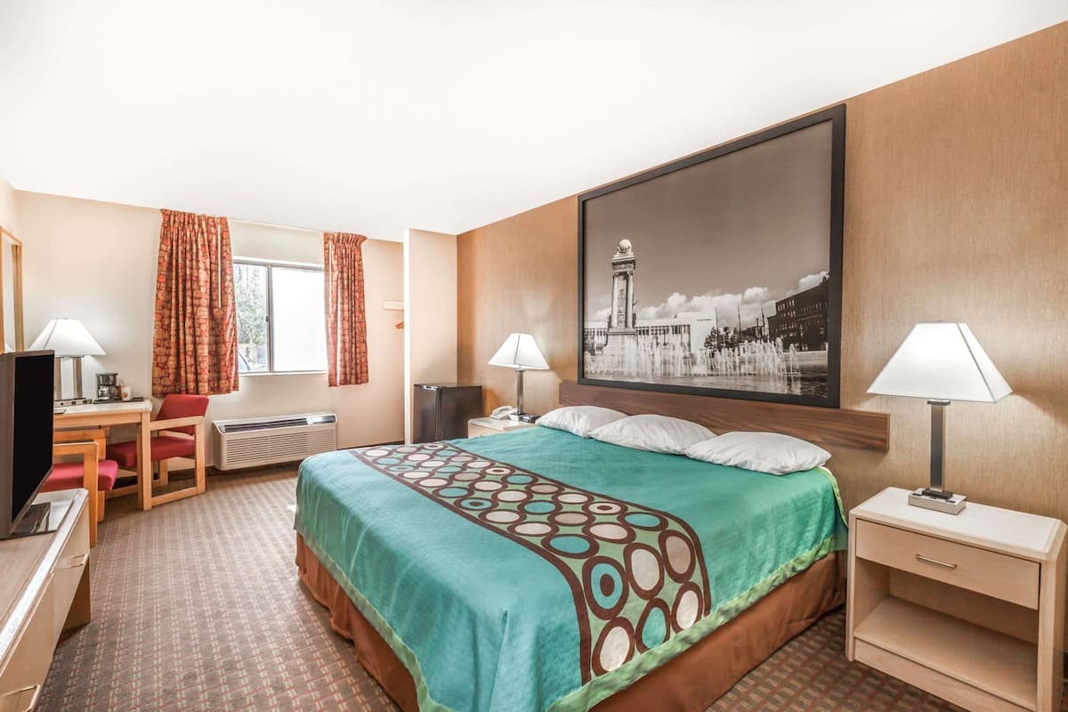 Ett hotellrum med en säng, ett skrivbord, en stol, en TV och ett stort fönster med gardiner.