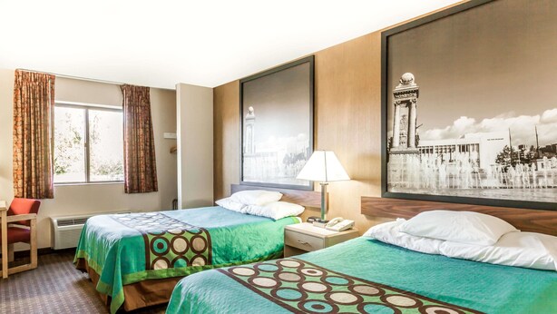 Habitación de hotel con dos camas, un escritorio, una silla, una mesita de noche con lámpara y un ventanal grande con cortinas.
