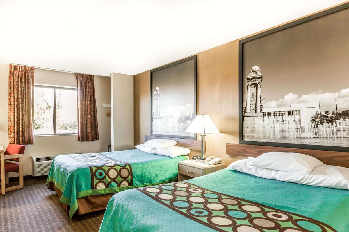 Ett hotellrum med två sängar, ett skrivbord, en stol, ett fönster med gardiner, en lampa och en stor tavla som föreställer en stadsbild.