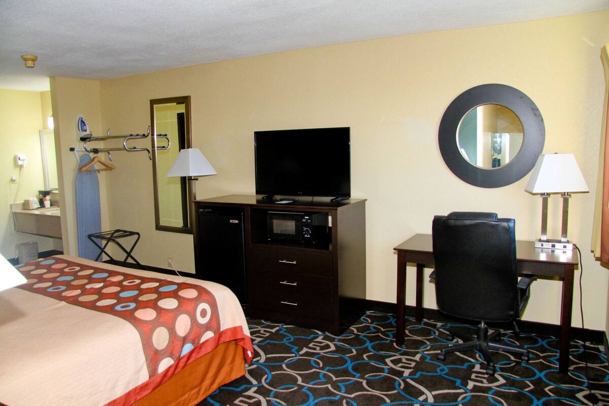 Habitación de hotel con cama, escritorio con silla, televisor, espejo y refrigerador pequeño.