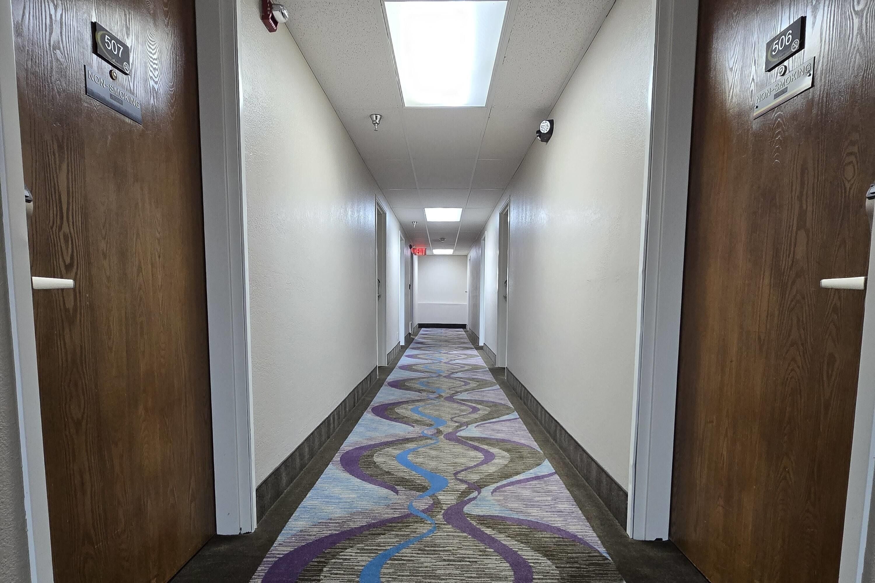 A hotel hallway with a patterned carpet, doors with room numbers, and a ceiling light.