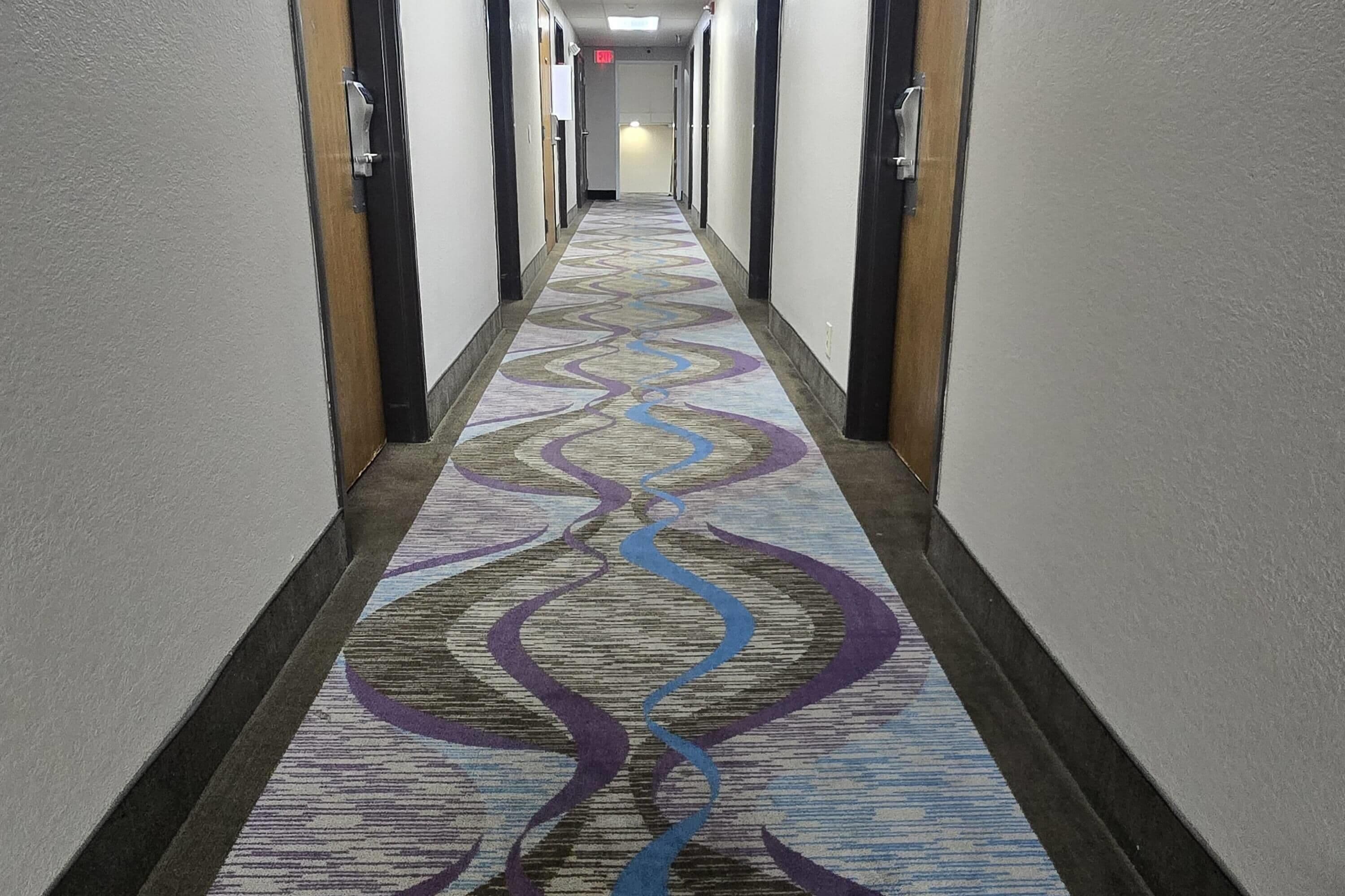 Un couloir d’hôtel avec un tapis à motifs, des portes de chaque côté et une lumière au bout.