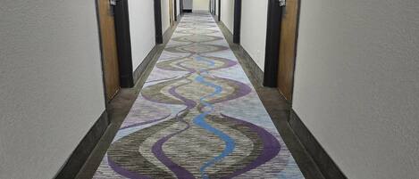 Un couloir d’hôtel avec un tapis à motifs, des portes de chaque côté et une lumière au bout.