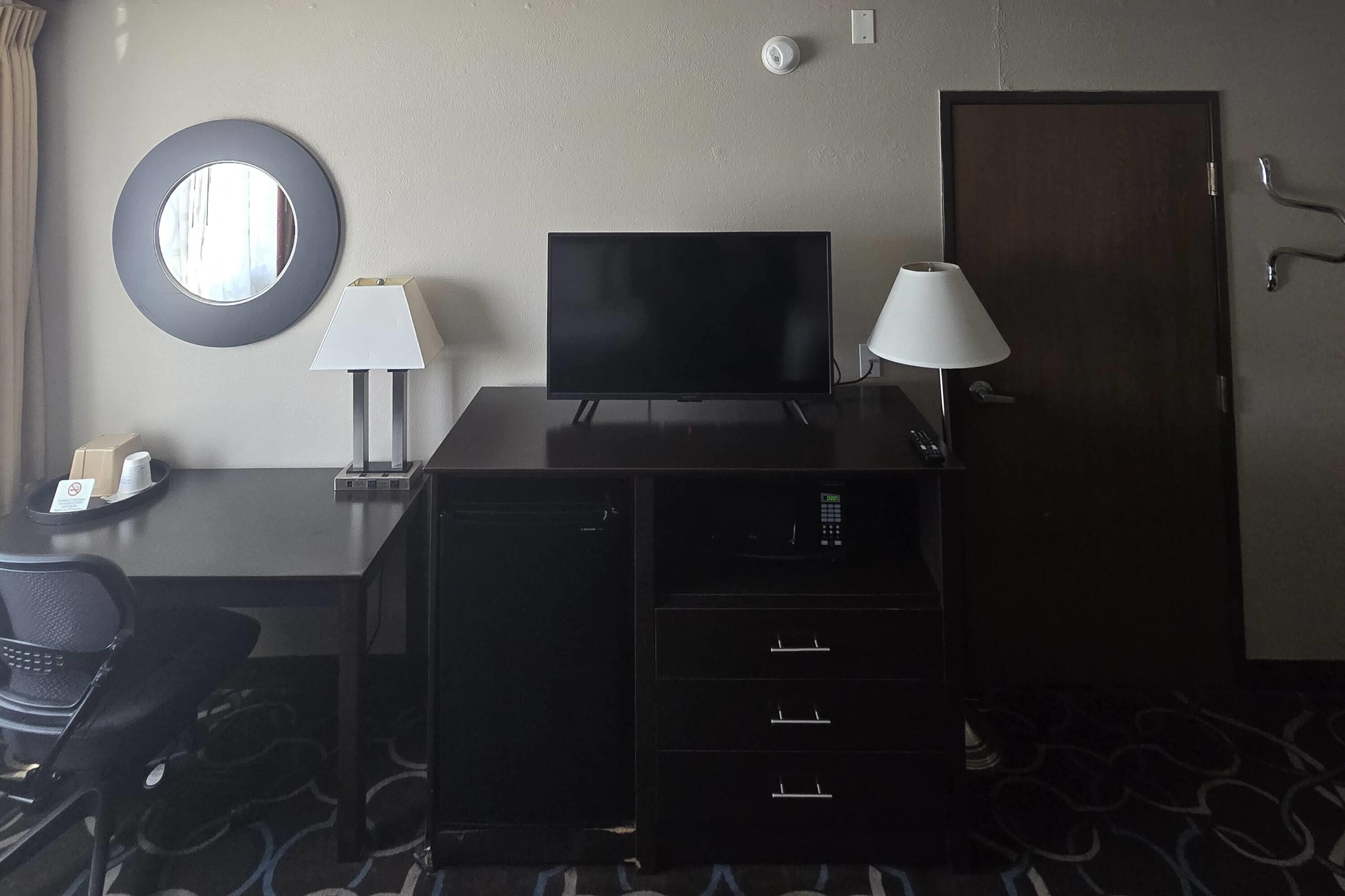 A hotel room with a flat-screen TV, two wall-mounted lamps, a desk with a chair, a coat rack, and a door.