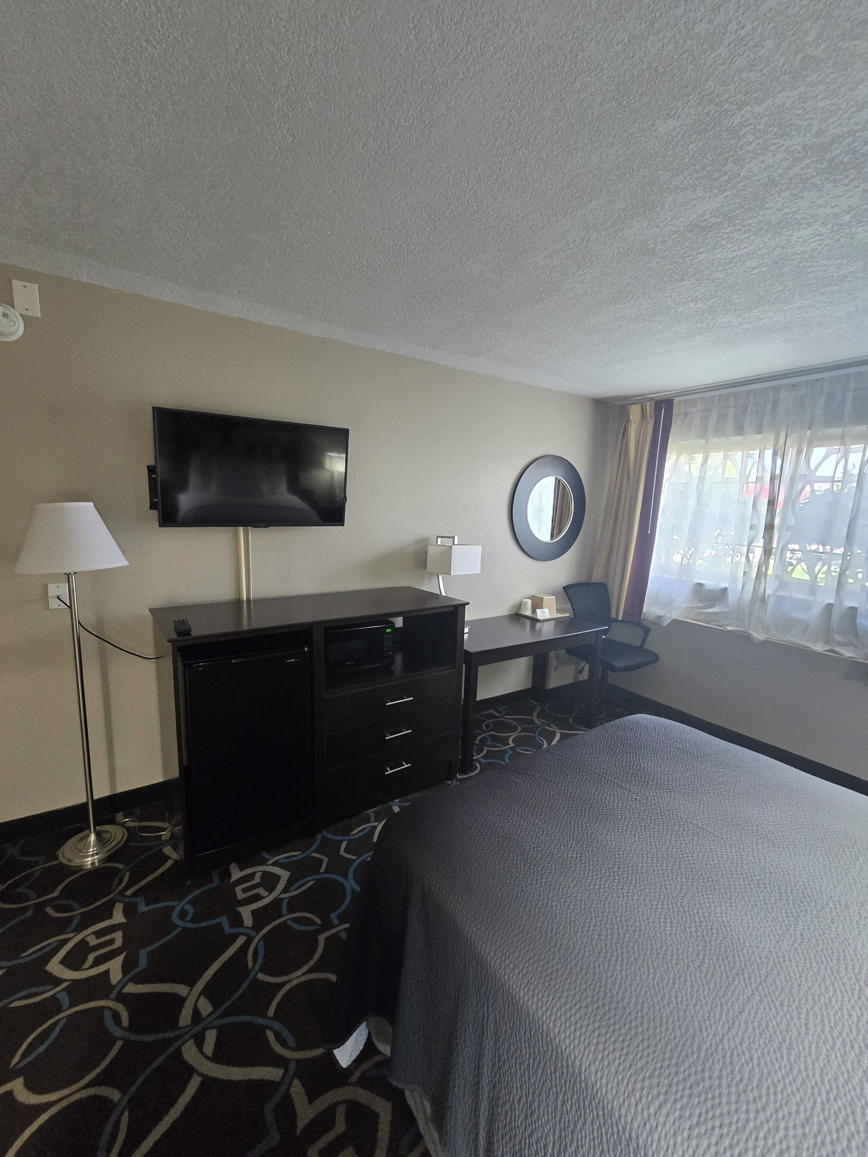 A hotel room with a bed, a desk, a chair, a flat-screen TV, a lamp, a mirror, and a window with curtains.
