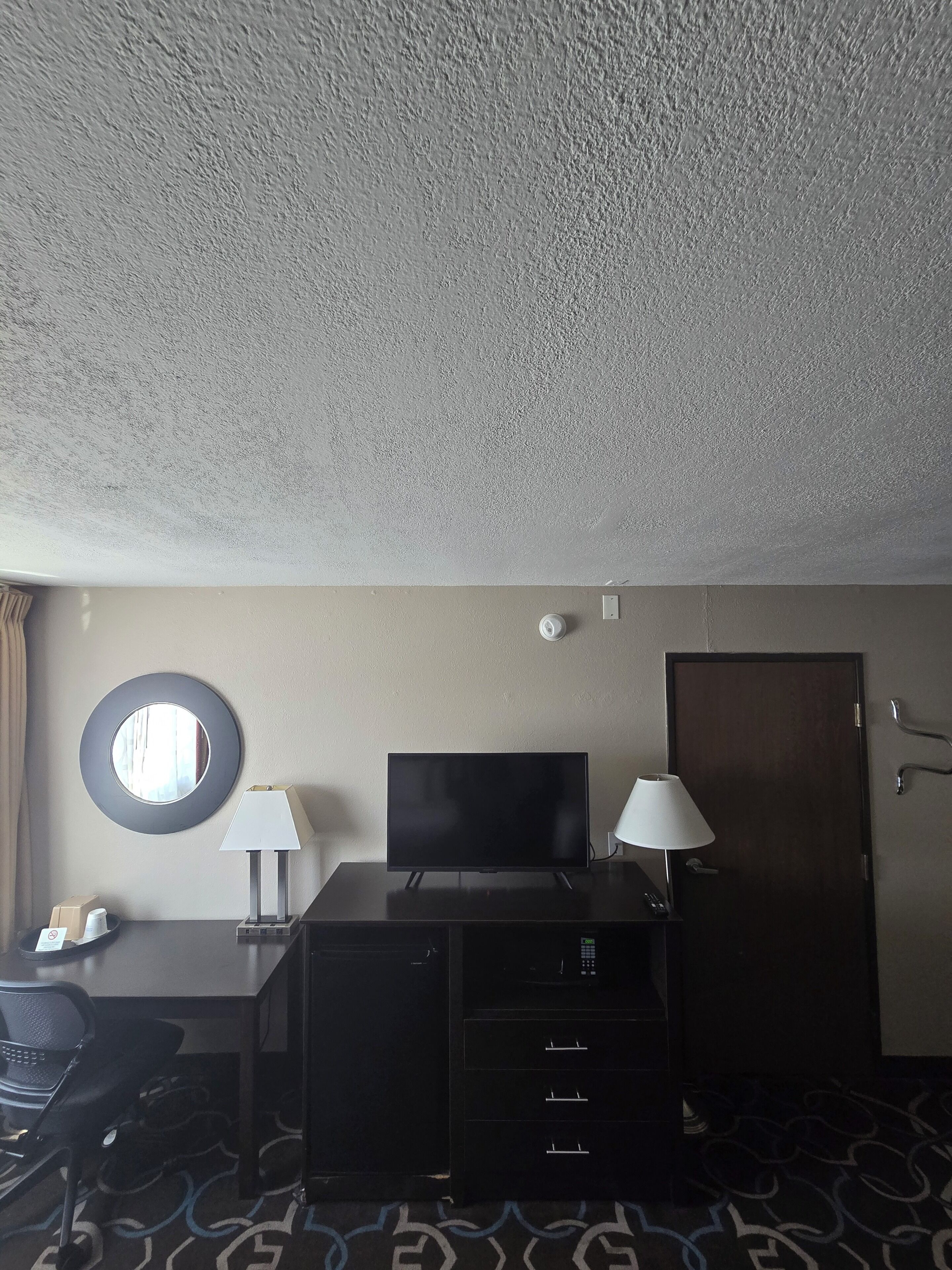 Een hotelkamer met een televisie, twee lampen, een bureau, een stoel, een ronde spiegel, een deur en een kamerbreed tapijt met een patroon.