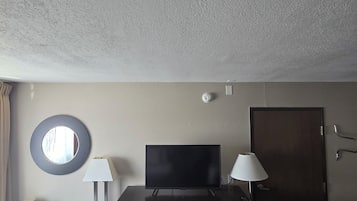 A hotel room with a television, two lamps, a desk, a chair, a circular mirror, a door, and a patterned carpet.