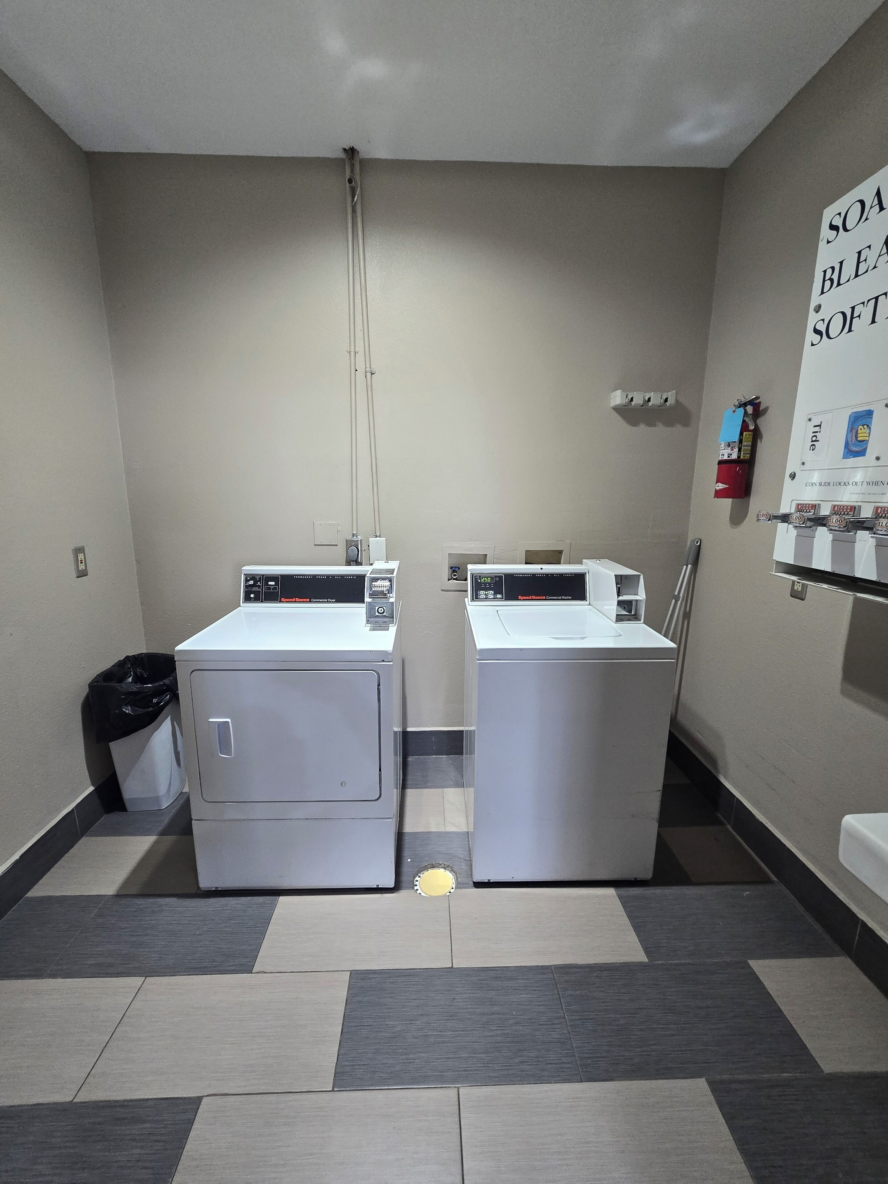 A laundry room with two front-loading washing machines and a dryer, a trash bin, and a fire extinguisher on the wall.